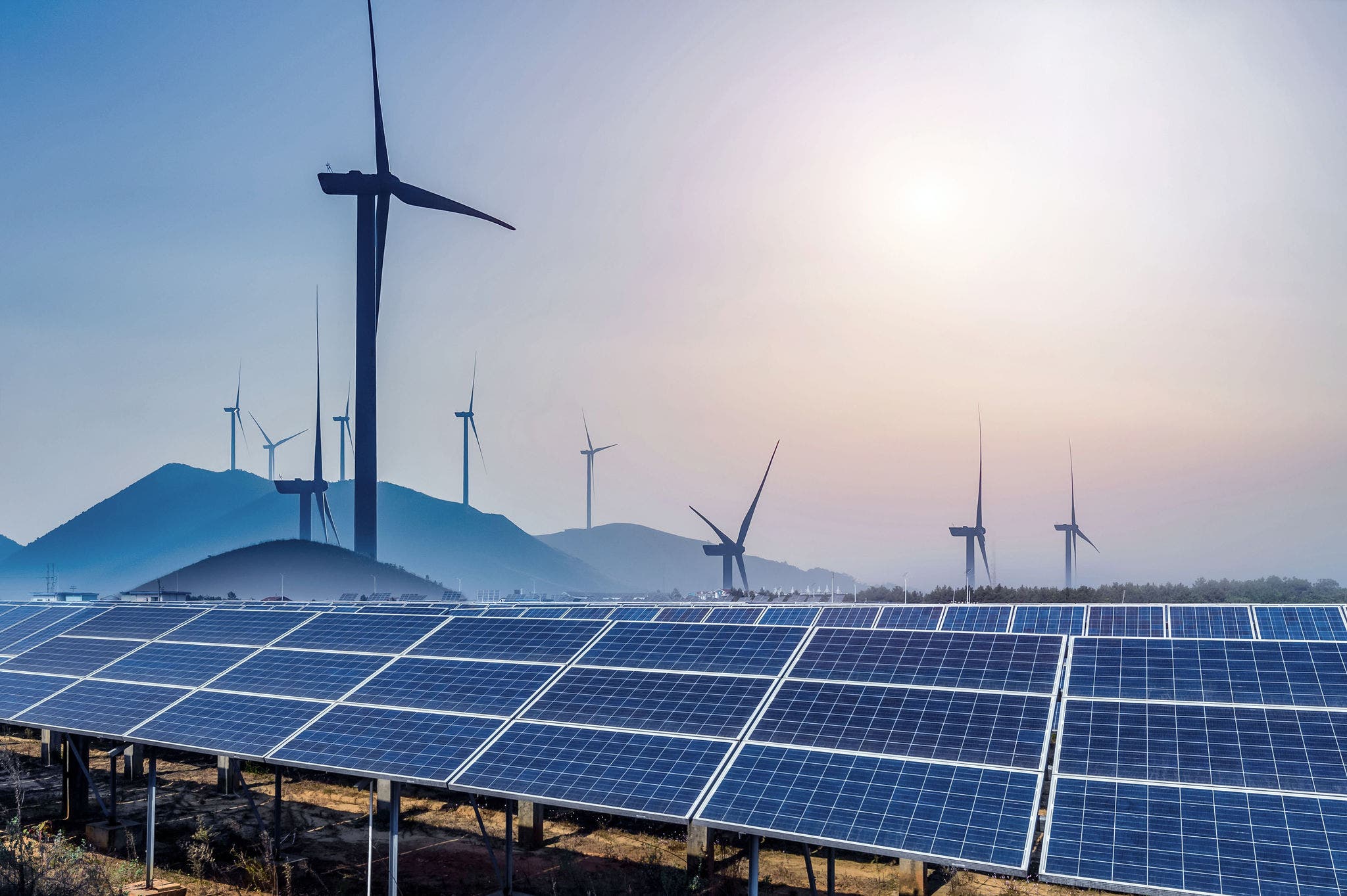 Solar panels in the foreground with multiple wind turbines in the background, set against a mountainous landscape under a clear sky, representing renewable energy sources.