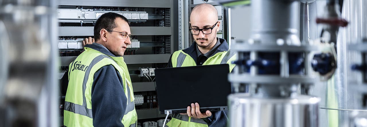 Two men in front of an electrical cabinet, a computer in the hands, working in production unit