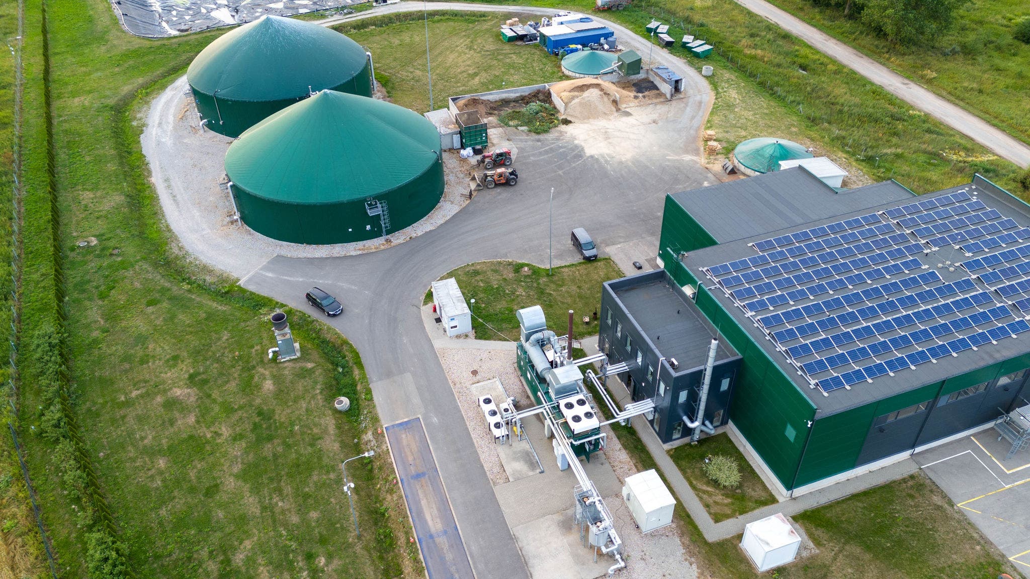 Aerial view of a modern biogas plant facility, featuring large green fermentation tanks and a solar-panel-covered building. The site includes storage areas and various processing equipment, highlighting its role in renewable energy production and sustainable practices. The image emphasizes clean energy solutions with keywords like biogas, solar power, and renewable energy.