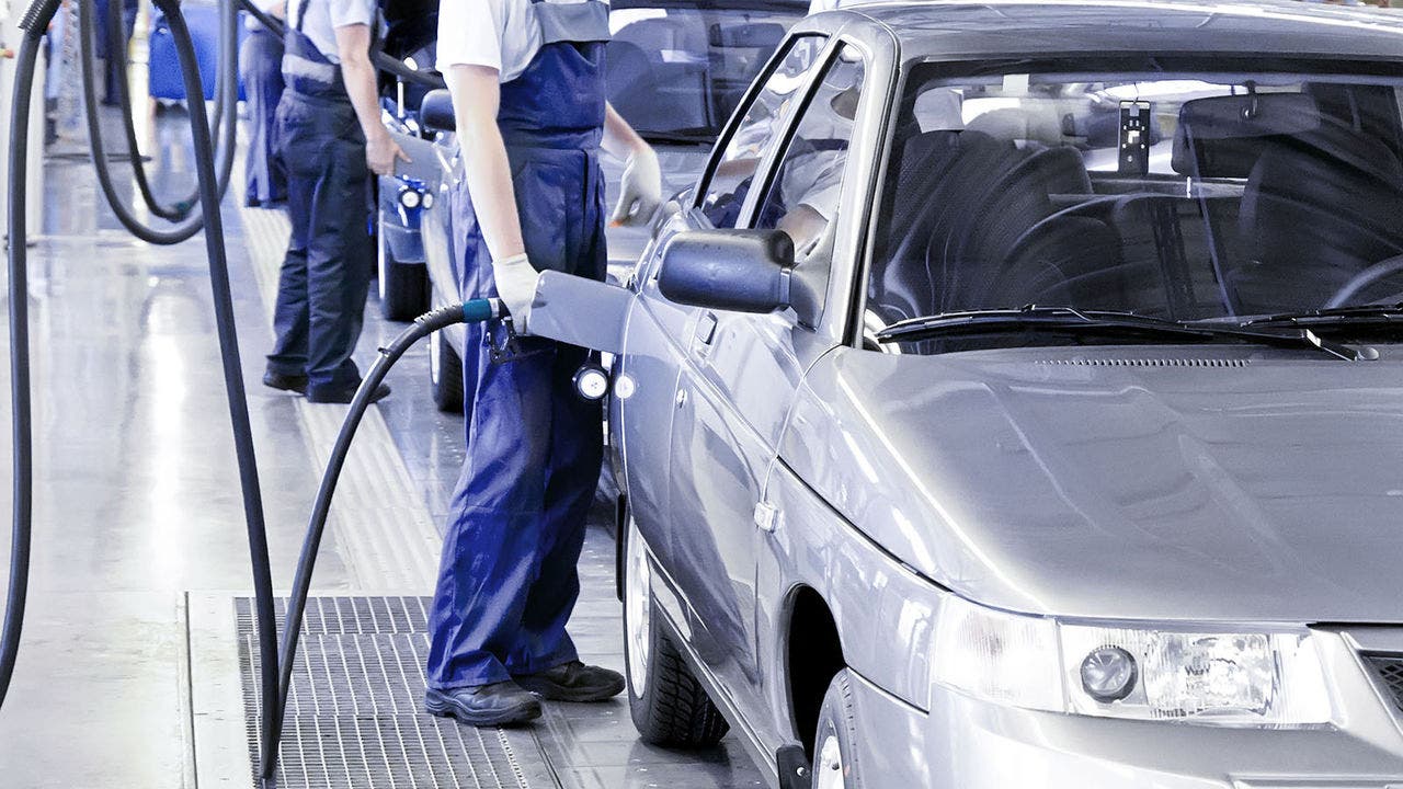 A worker in a blue jumpsuit is standing next to a silver car in an automotive manufacturing facility, holding a hose connected to the vehicle
