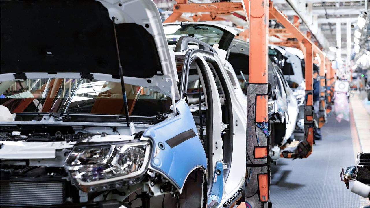 Partially assembled car bodies on an assembly line in an automotive factory, with hoods open and supported by orange structures, highlighting component installation stage.