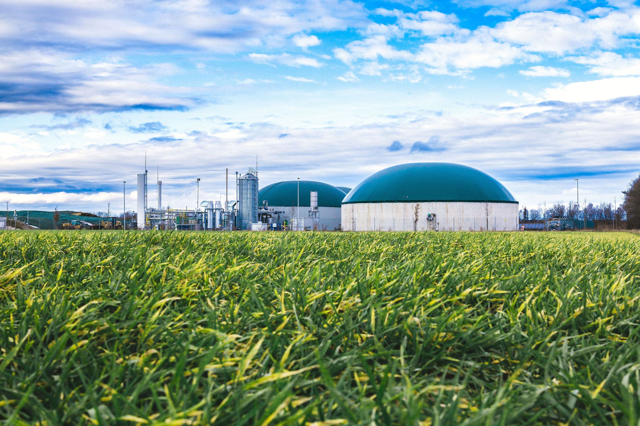 Biogas plant with green storage domes situated in a lush grassy field under a partly cloudy blue sky.
