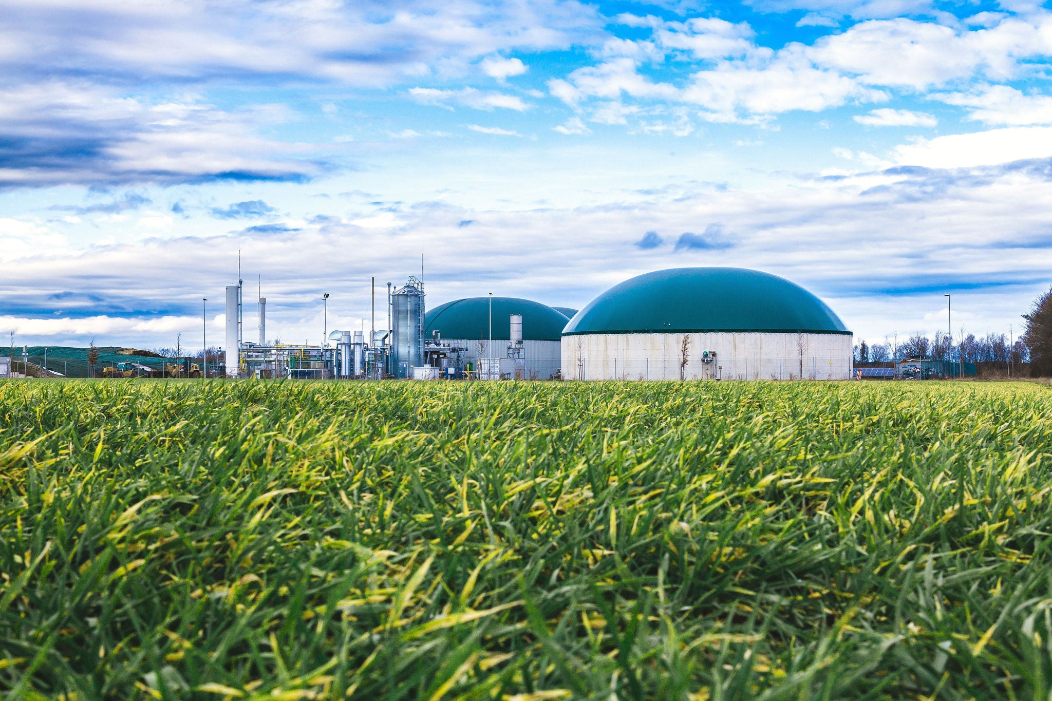 Biogas plant with green storage domes situated in a lush grassy field under a partly cloudy blue sky.
