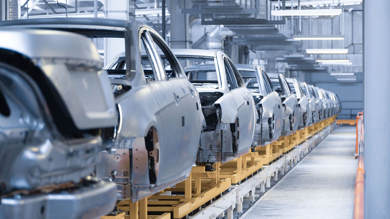 A row of unfinished car bodies is lined up in an automotive manufacturing assembly line. The vehicles are positioned on yellow platforms in a spacious, well-lit industrial facility, highlighting the manufacturing process before components are added.