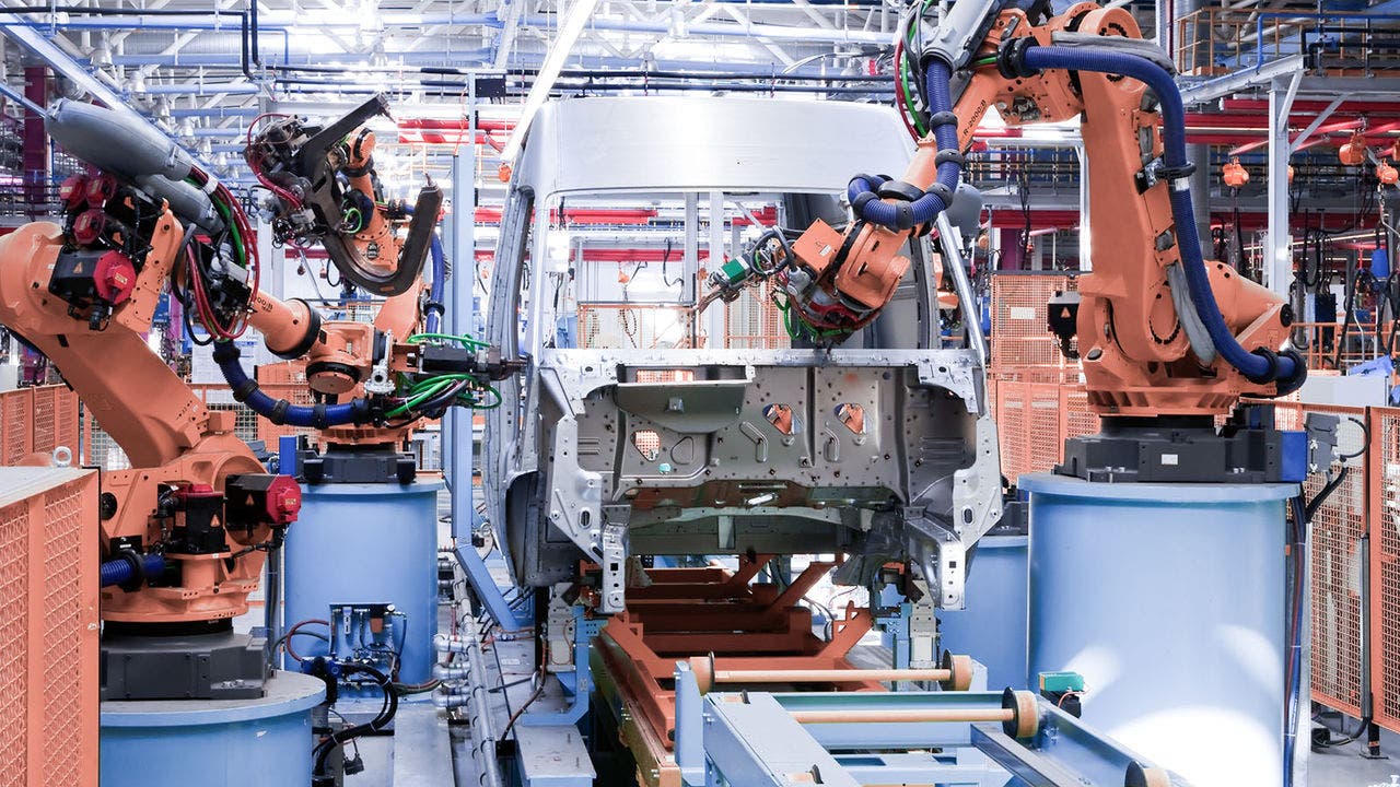 An array of orange robotic arms in a factory setting assembling a vehicle chassis on a production line